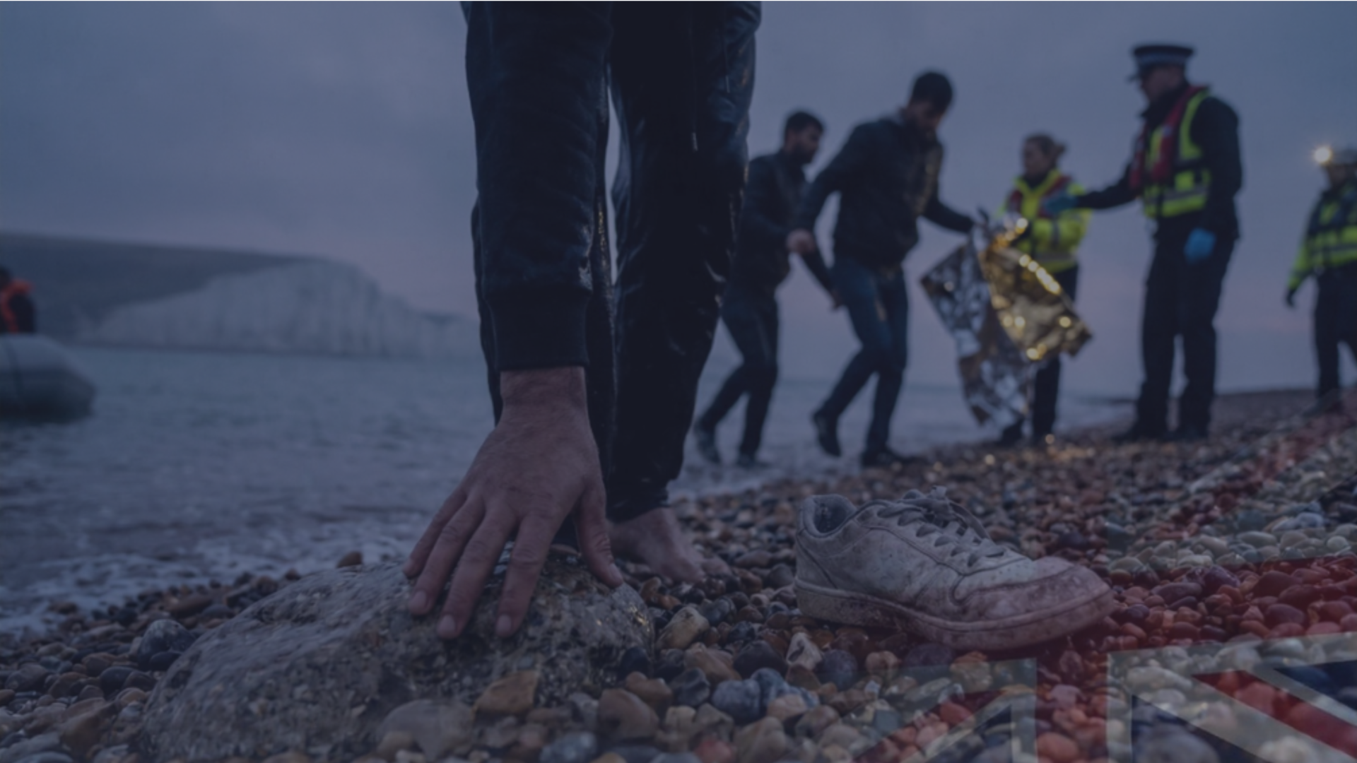 A close-up photograph of a bare hand touching a large, flat stone on a shingle beach, with a discarded white sneaker lying nearby. In the background, a line of people walks up from the sea, toward a group of individuals in official high-visibility jackets and a helmeted responder with a headlamp. Prominent white cliffs are visible on the far horizon under a dark, overcast sky.